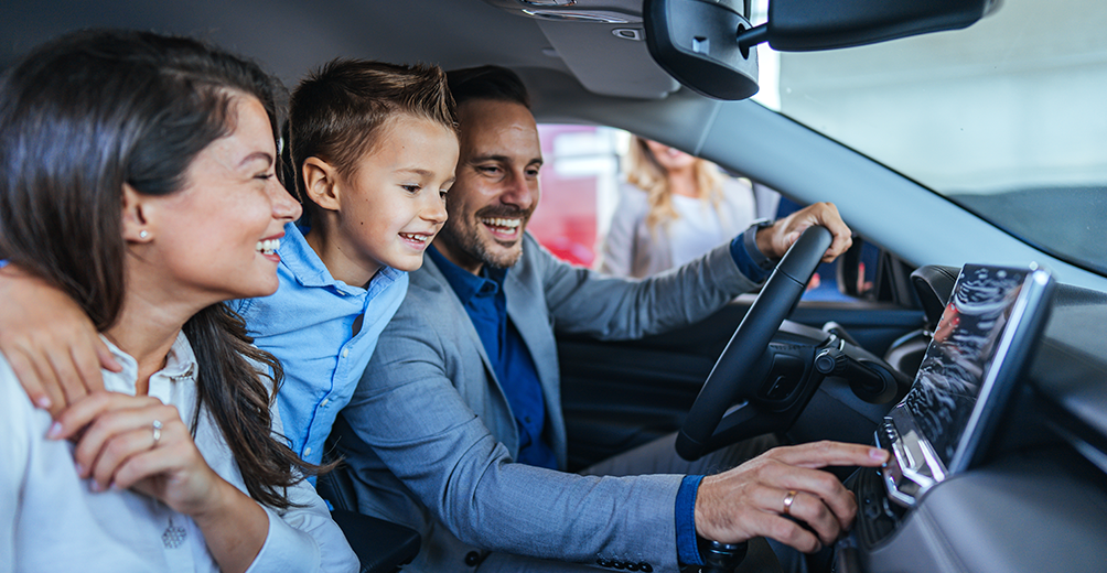 Família animada de três sentada dentro de um carro, a explorar as características interiores do veículo. O pai está a mostrar as funções do painel enquanto a mãe e o filho olham felizes.